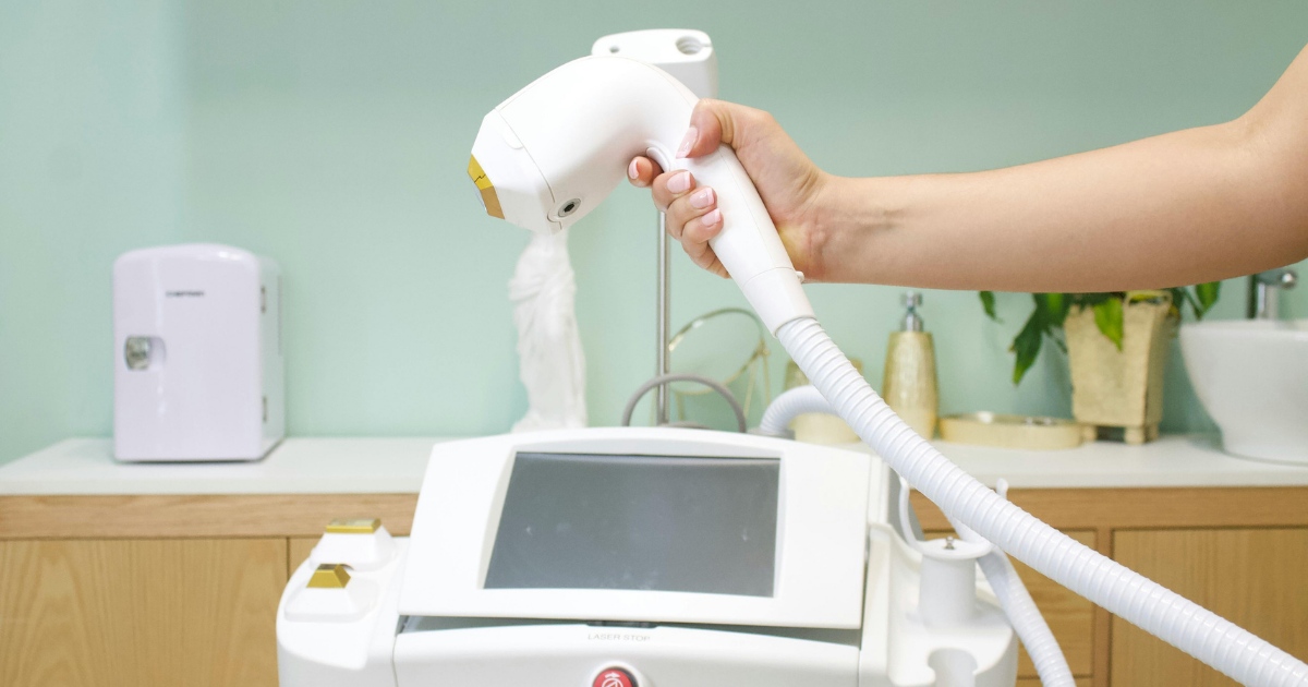 “Close-up of a handheld IPL device held above a clinical treatment machine, with a touch-screen console and hose attachment, in a clean, modern treatment room.