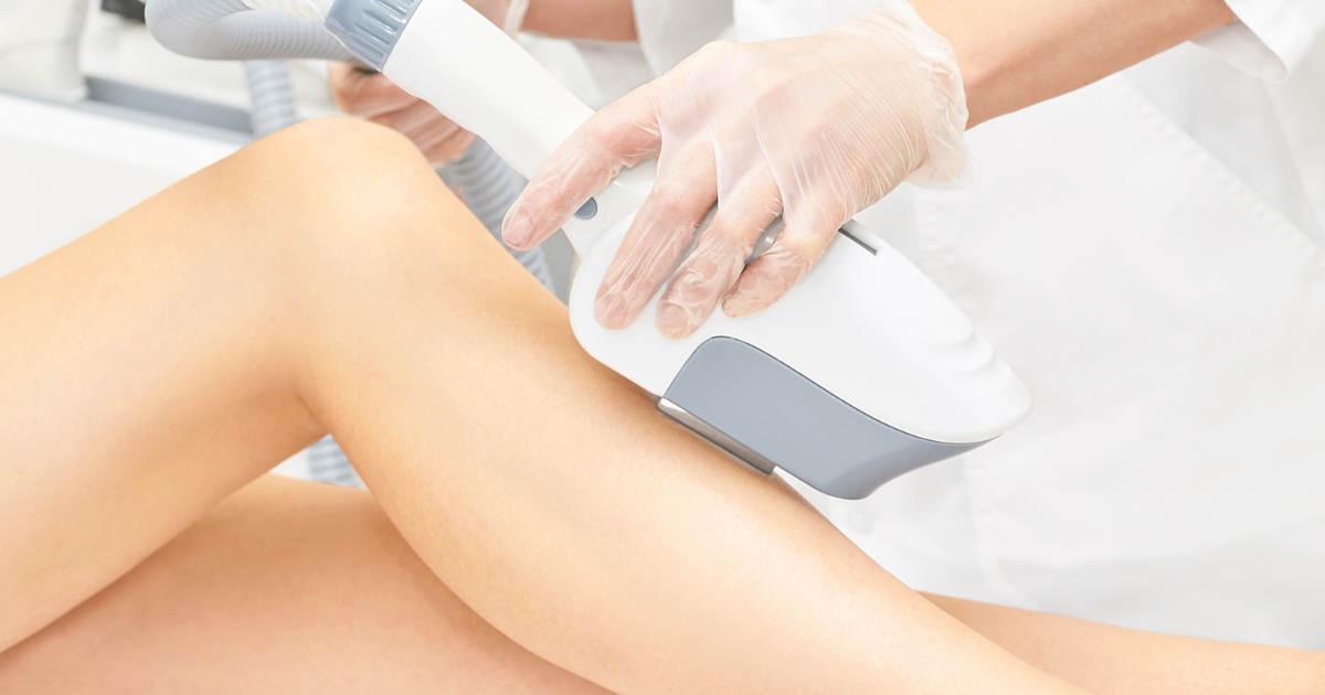 Close-up of a cosmetic treatment in progress, with a practitioner wearing gloves using a handheld body-contouring device on a person’s thigh in a clean clinical setting.