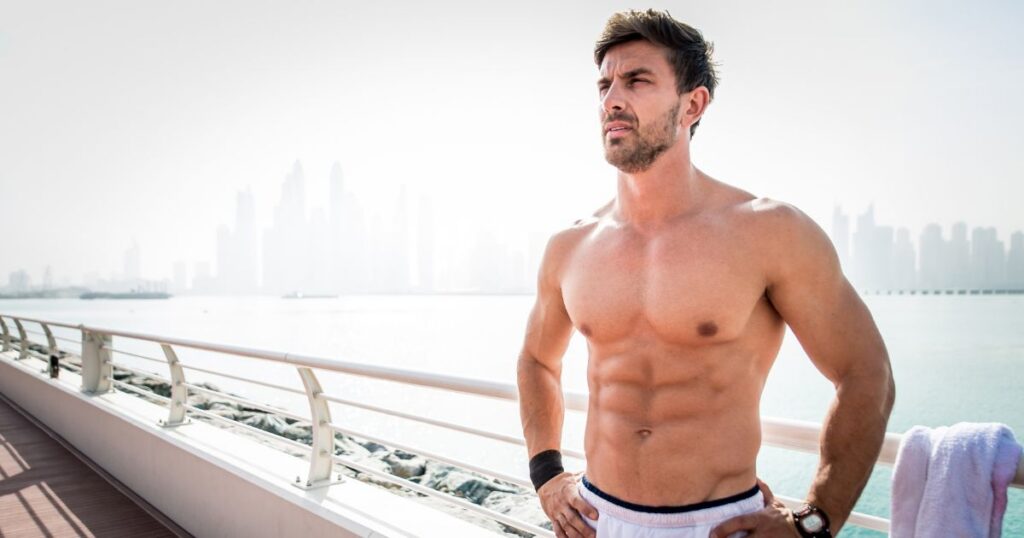 Shirtless fit man standing by a waterfront railing with a city skyline in the background.